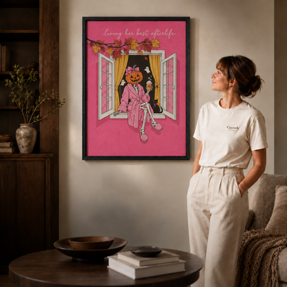 Woman in a white tee admiring a pink pumpkin skeleton Halloween canvas with autumn leaves in a black frame.