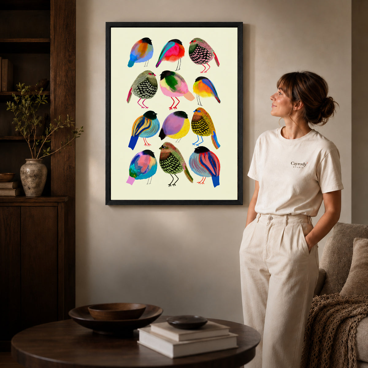 Colorful birds canvas art in a black frame above stacked books in a cozy living room.