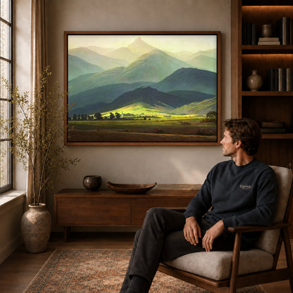 Mountain valley canvas art above a wooden console near a seated man and leafy branches.