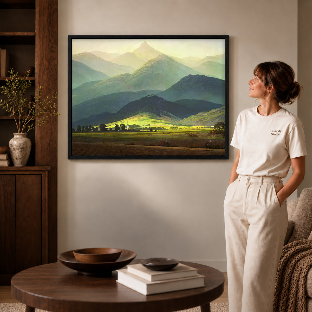 Mountain valley canvas art in a black frame above stacked books in a cozy living room.