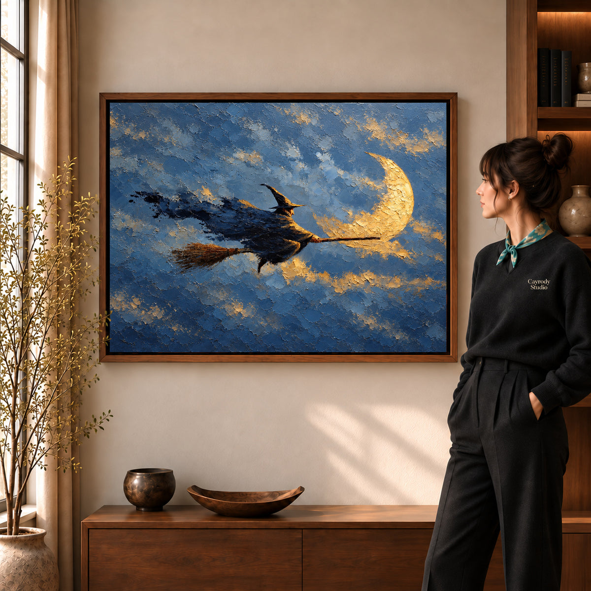 Moonlit witch canvas art in a walnut frame beside a woman in a warmly lit living room.