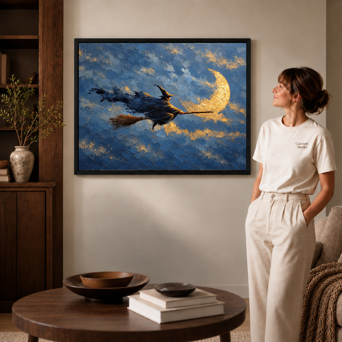 Moonlit witch canvas art in a black frame above stacked books in a cozy living room.