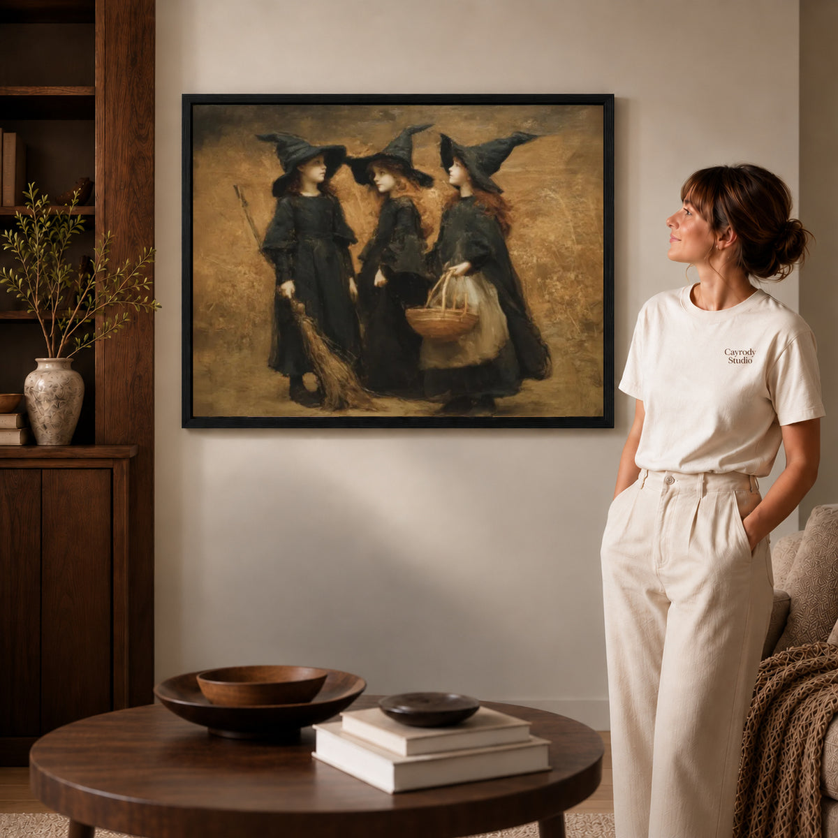 Three witches canvas art displayed in a black frame beside a woman, with soft room lighting.