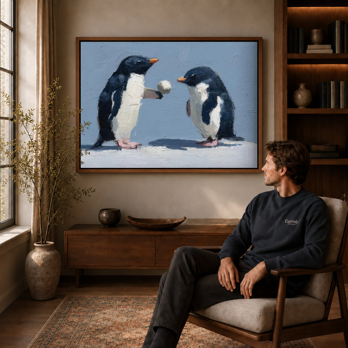 Penguin pair canvas art above a wooden console near a seated man and leafy branches.