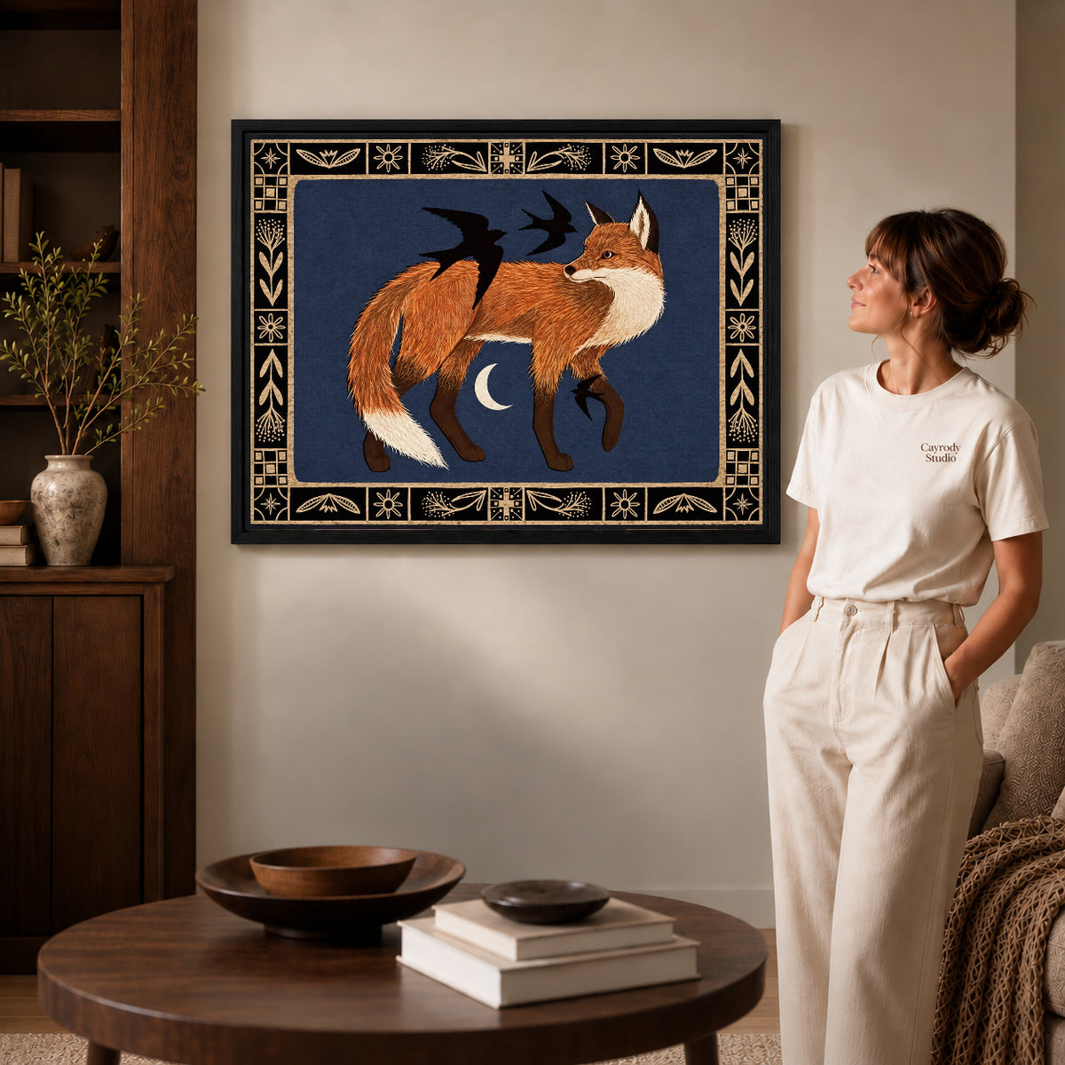 Fox and birds canvas art in a black frame above stacked books in a cozy living room.