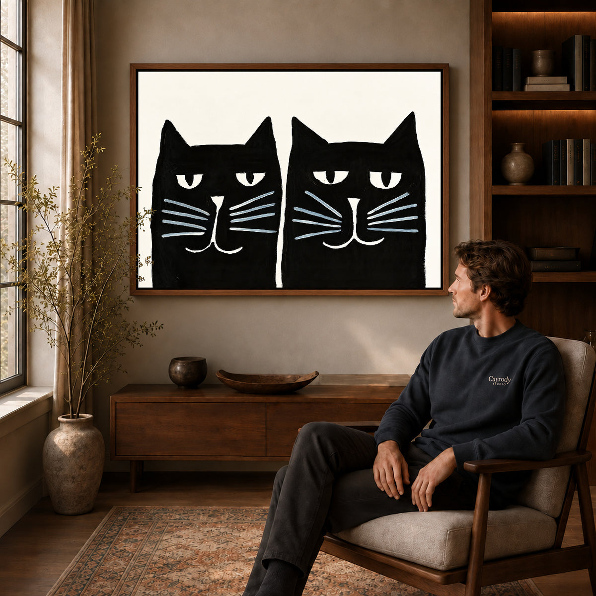 Black cat pair canvas art above a wooden console near a seated man and leafy branches.