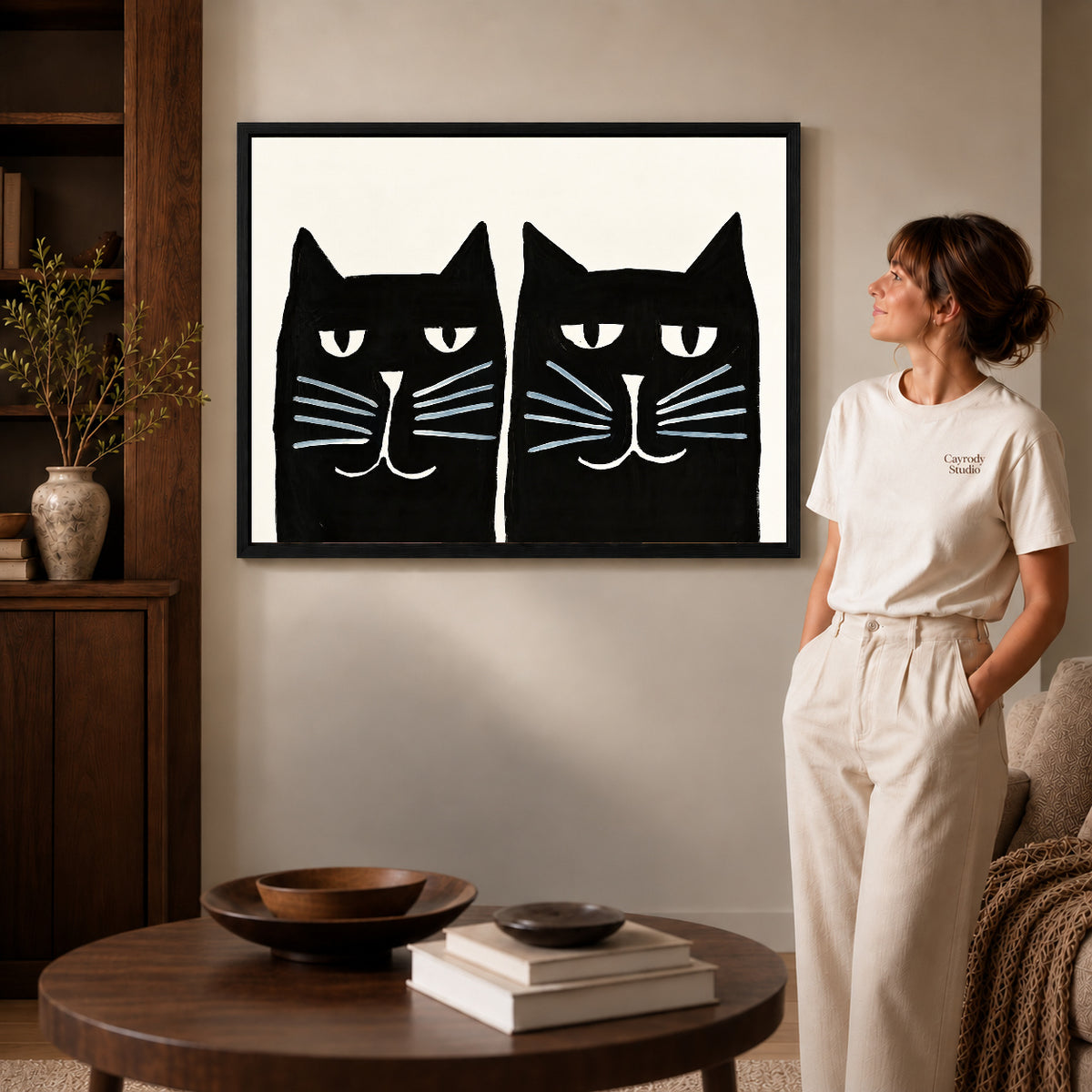 Black cat pair canvas art in a black frame above stacked books in a cozy living room.