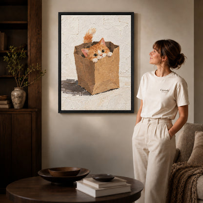 Paper bag kitten canvas art in a black frame above stacked books in a cozy living room.