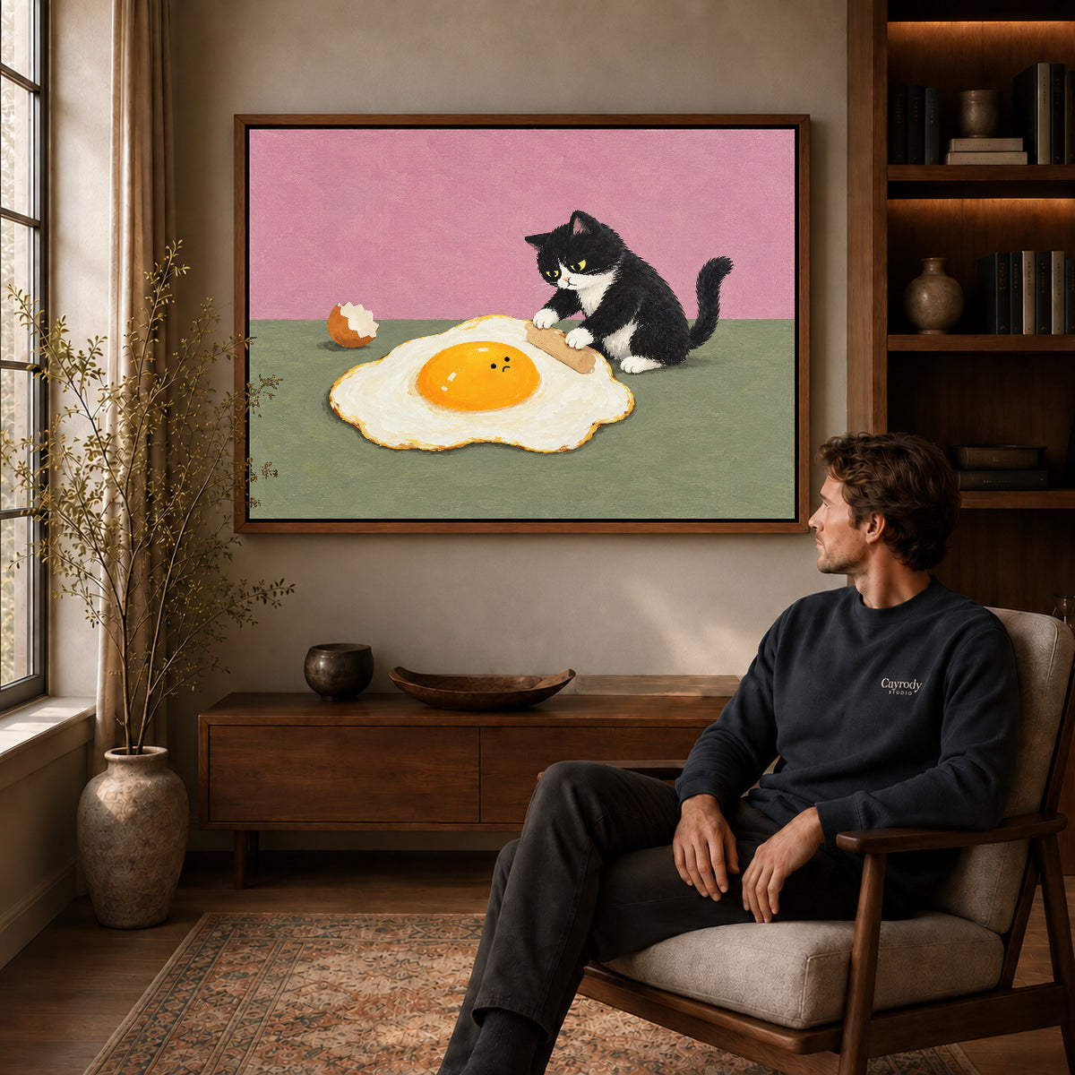 playful egg rolling cat canvas art in a walnut frame above a console beside a seated man.