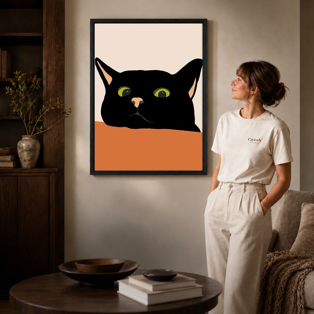 Black cat canvas art in a black frame above stacked books in a cozy living room.
