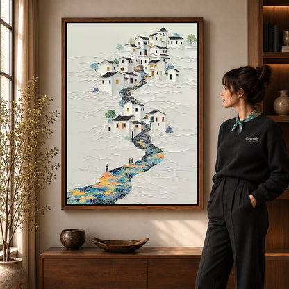 Hillside village canvas art in a walnut frame beside a woman in a warmly lit living room.