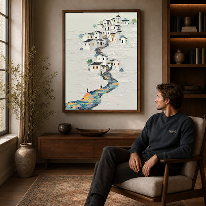 Hillside village canvas art above a wooden console near a seated man and leafy branches.