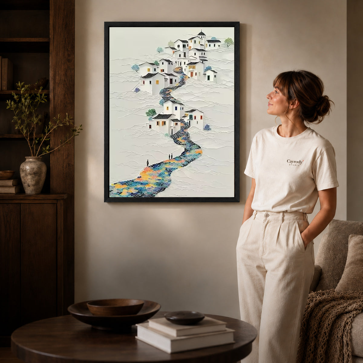 Hillside village canvas art in a black frame above stacked books in a cozy living room.