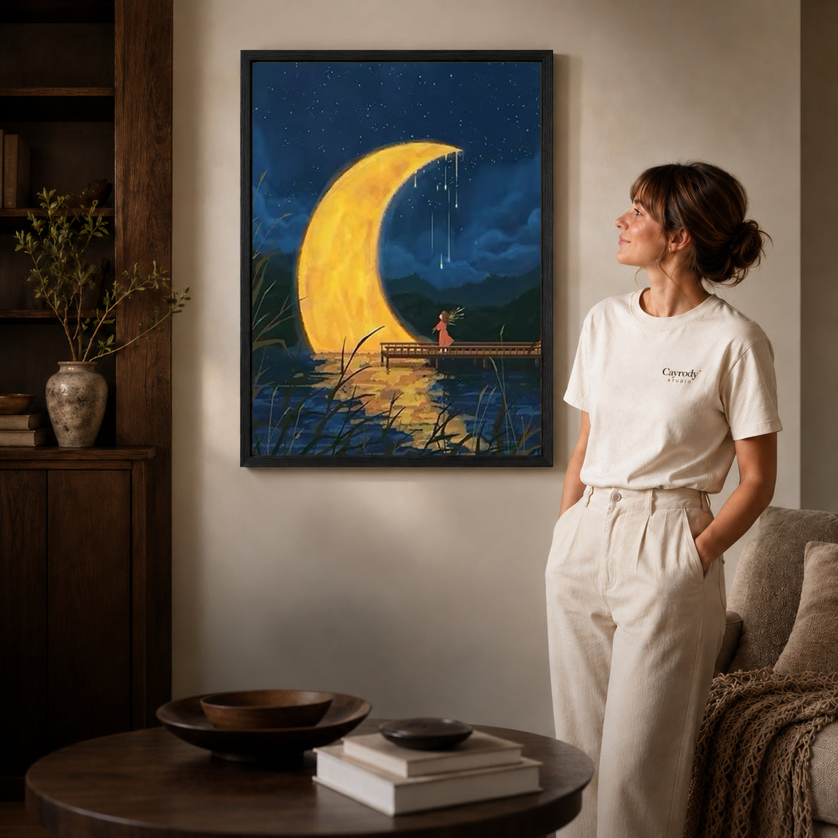 Crescent moon canvas art in a black frame above stacked books in a cozy living room.