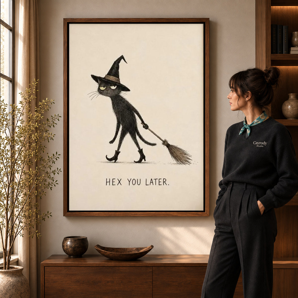 Broomstick Cat canvas art in a walnut frame beside a standing woman and wooden console.
