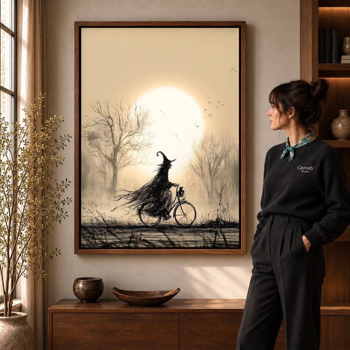 Cycling Witch canvas art in a walnut frame beside a standing woman and wooden console.