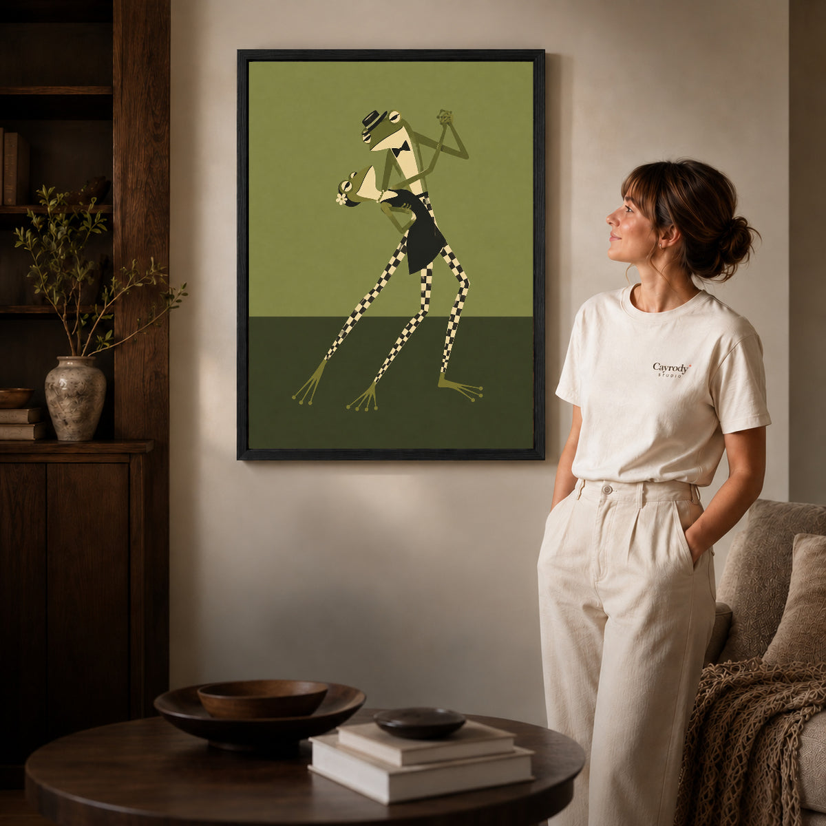 Black framed dancing frogs canvas art displayed beside a woman in a warm living room.