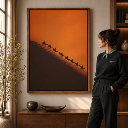 Desert Caravan canvas art in a walnut frame beside a standing woman and wooden console.