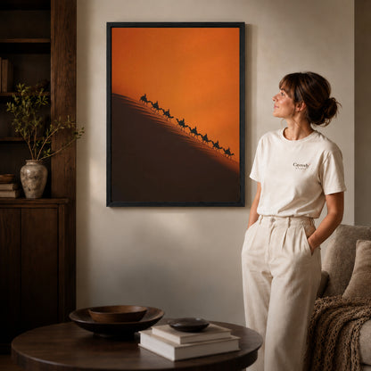 Black framed desert caravan canvas art displayed beside a woman in a warm living room.