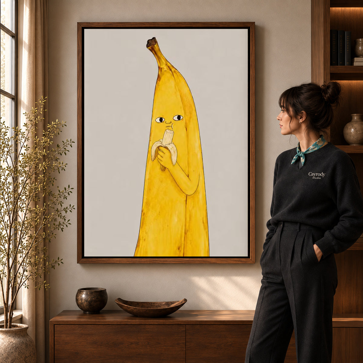 Banana Snack canvas art in a walnut frame beside a standing woman and wooden console.