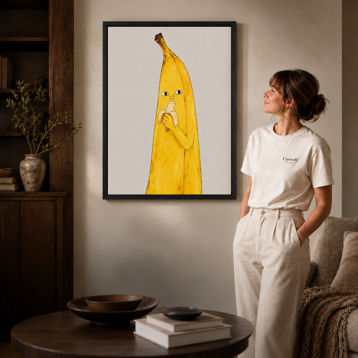 Banana Snack canvas art in a black frame above books in a welcoming living room.