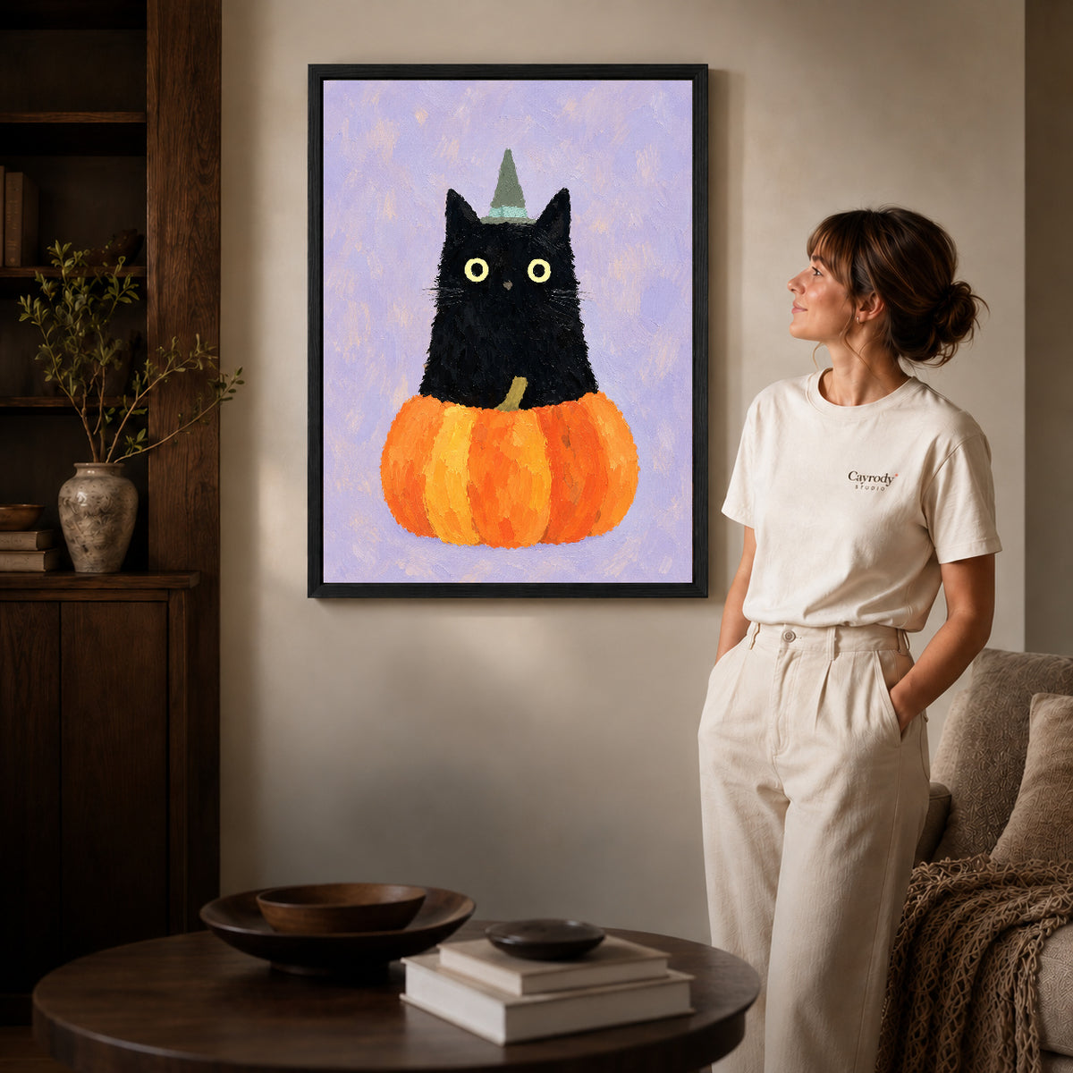 Black framed pumpkin witch cat canvas art displayed beside a woman with natural light across the wall.