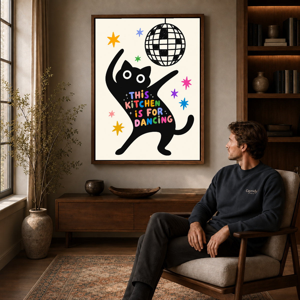 dancing kitchen cat canvas art in a walnut frame above a console beside a seated man.