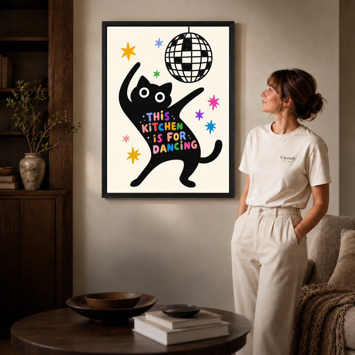dancing kitchen cat canvas art in a black frame beside a woman and round wooden table.