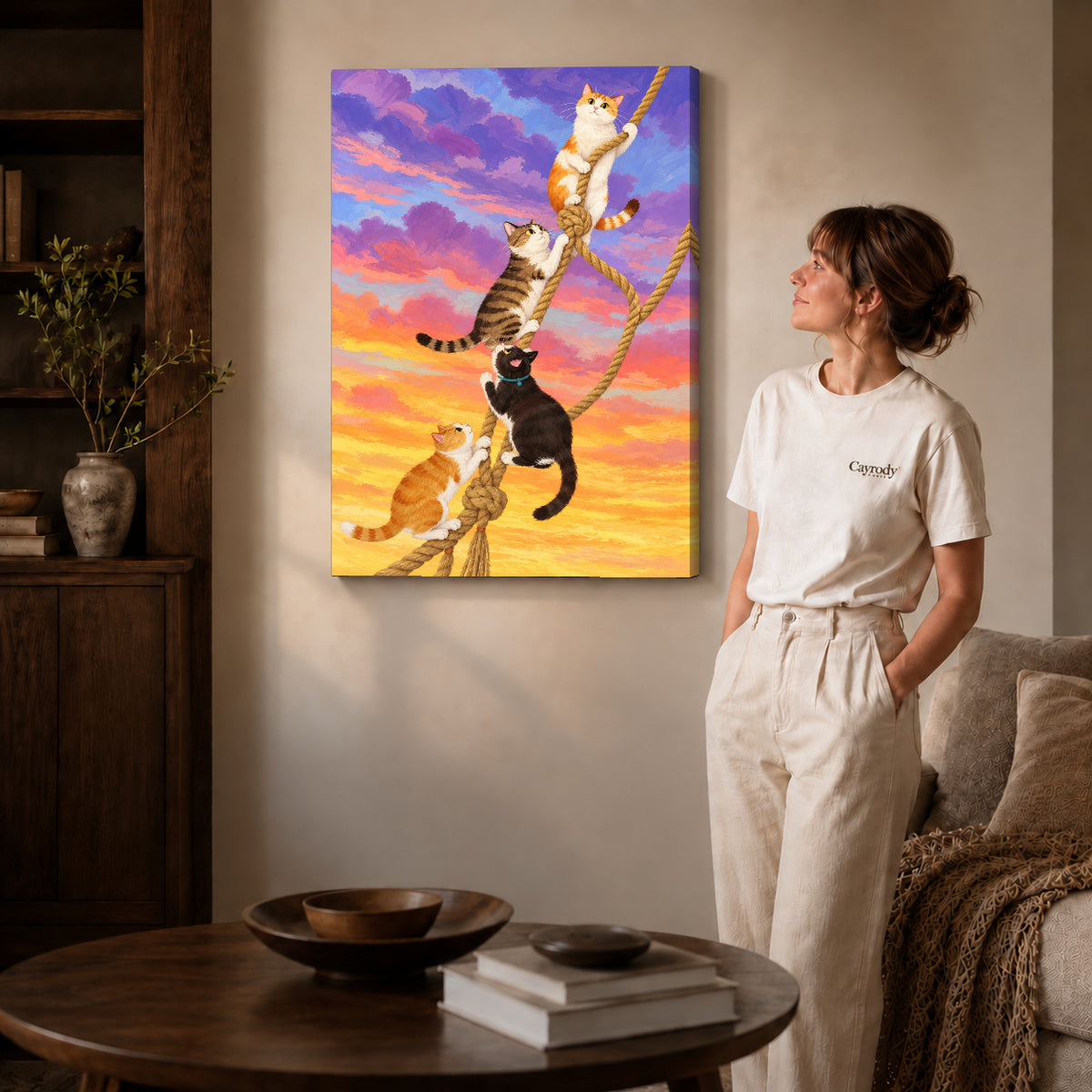 Gallery wrapped whimsical climbing cats canvas art with subtly shaded canvas edges beside a woman wearing cream clothing.