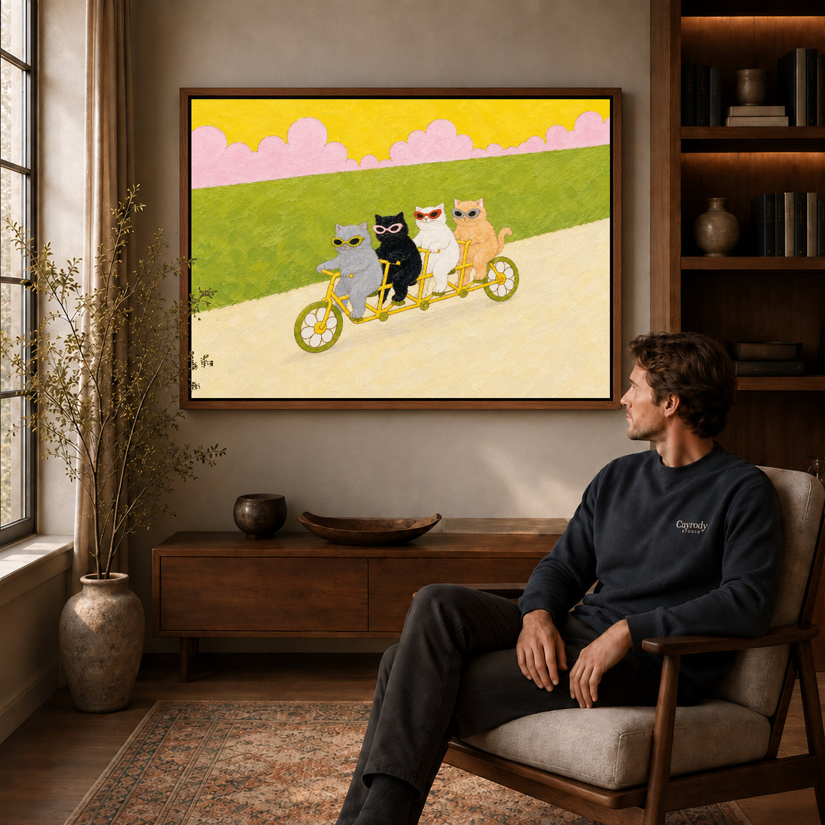 Walnut framed landscape tandem cycling cats canvas art above a seated man in a cozy wood furnished living room.