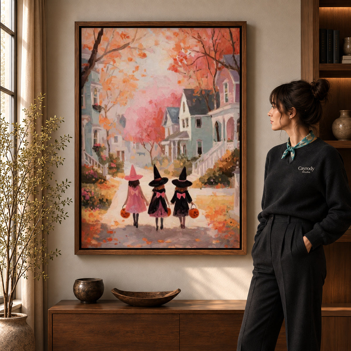 Little witches canvas art in a walnut frame beside a standing woman and wooden console.