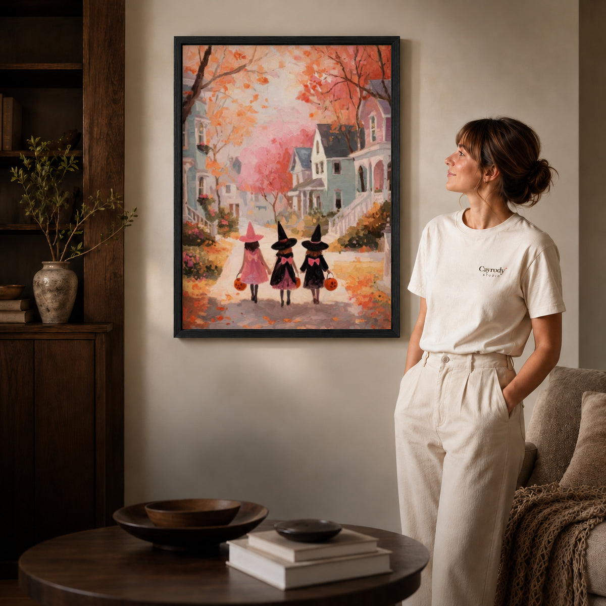 Little witches canvas art in a black frame beside a woman and round wooden table.