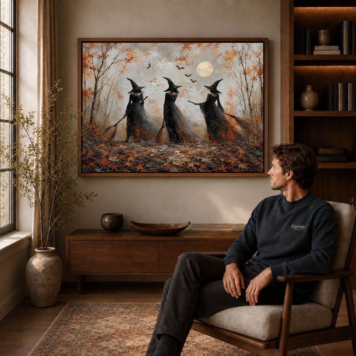 Woodland witches canvas art in a walnut frame above a console beside a seated man.