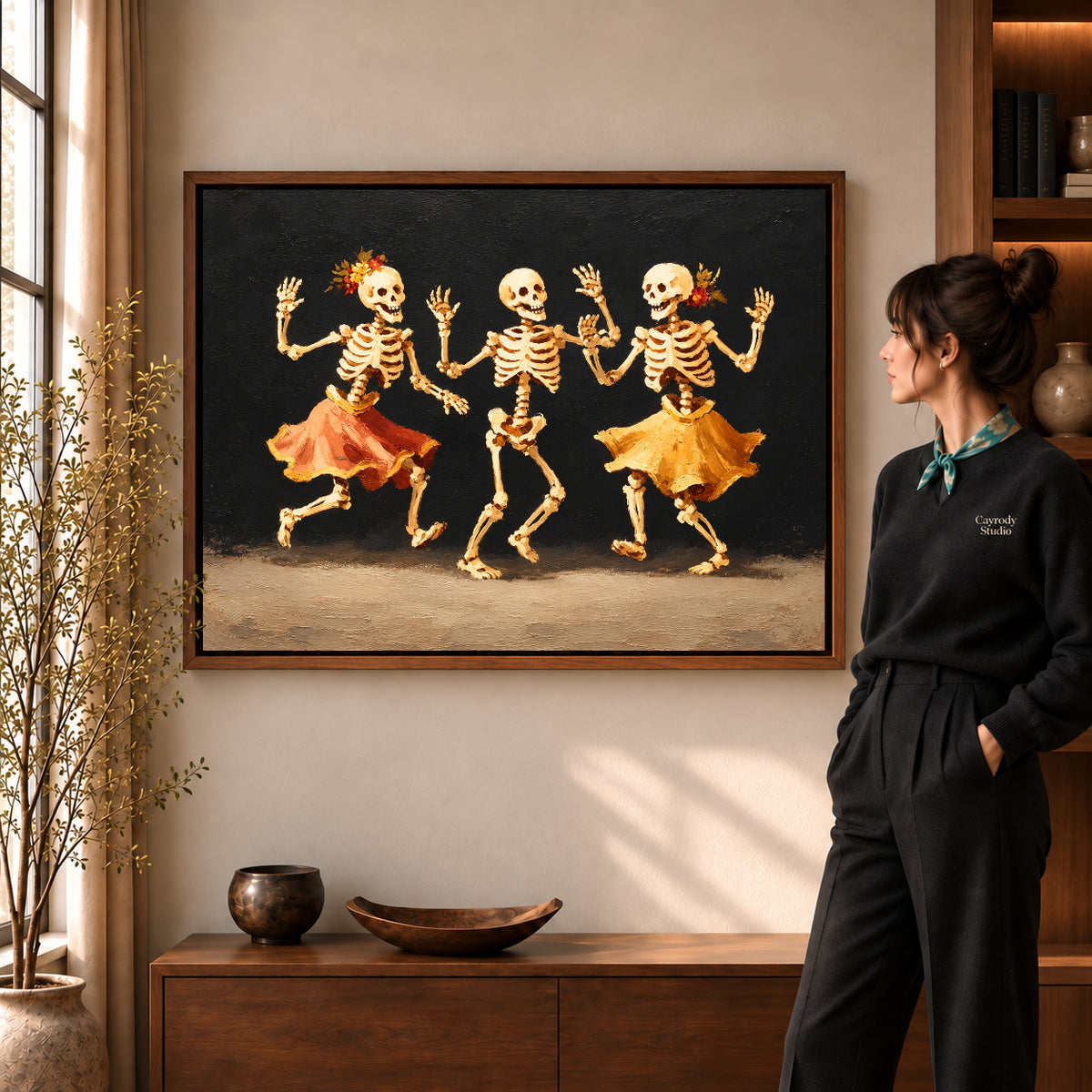 Dancing Skeletons canvas art in a walnut frame beside a standing woman and wooden console.