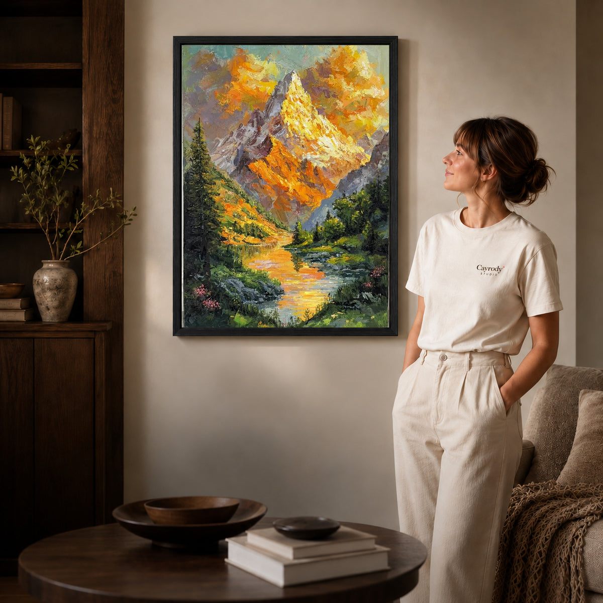 Alpine lake canvas art in a black frame beside a woman and round wooden table.