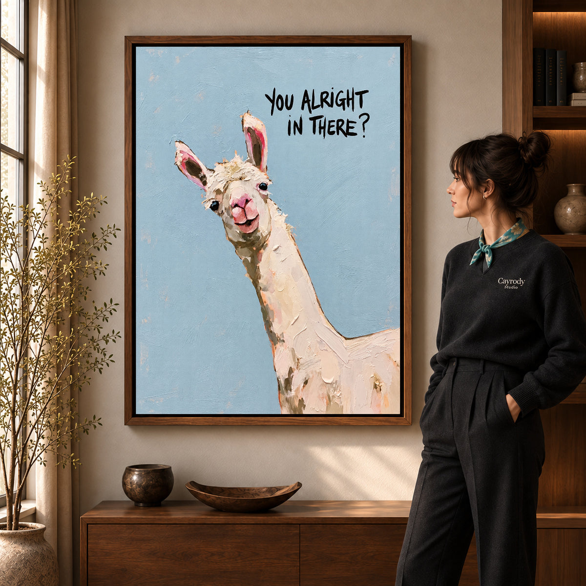 Curious llama canvas art in a walnut frame beside a standing woman and wooden console.