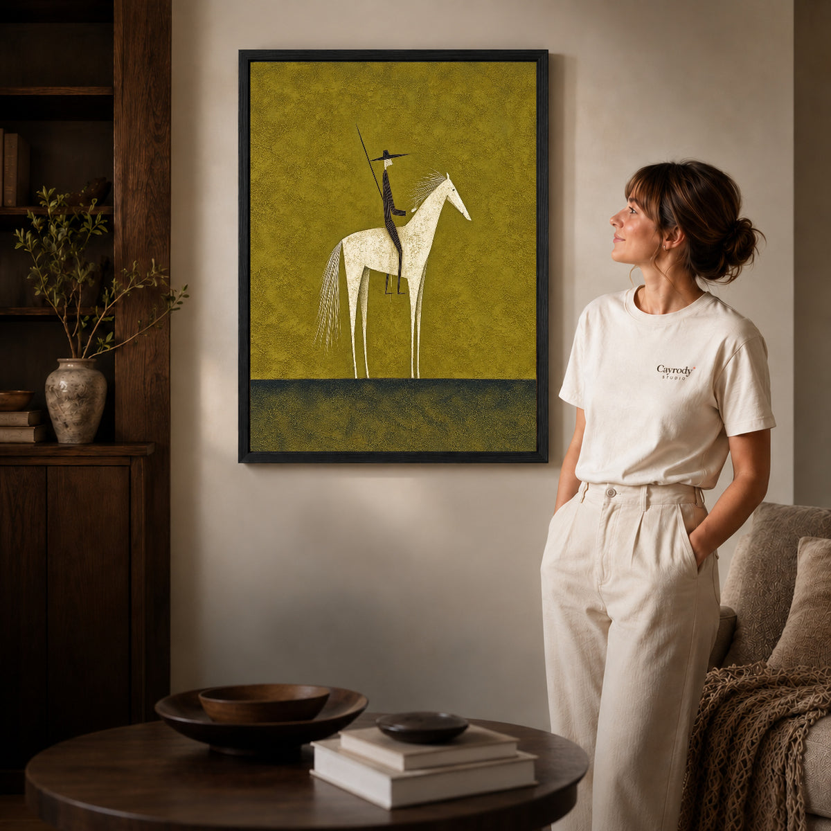 Folk horse canvas art in a black frame beside a woman and round wooden table.