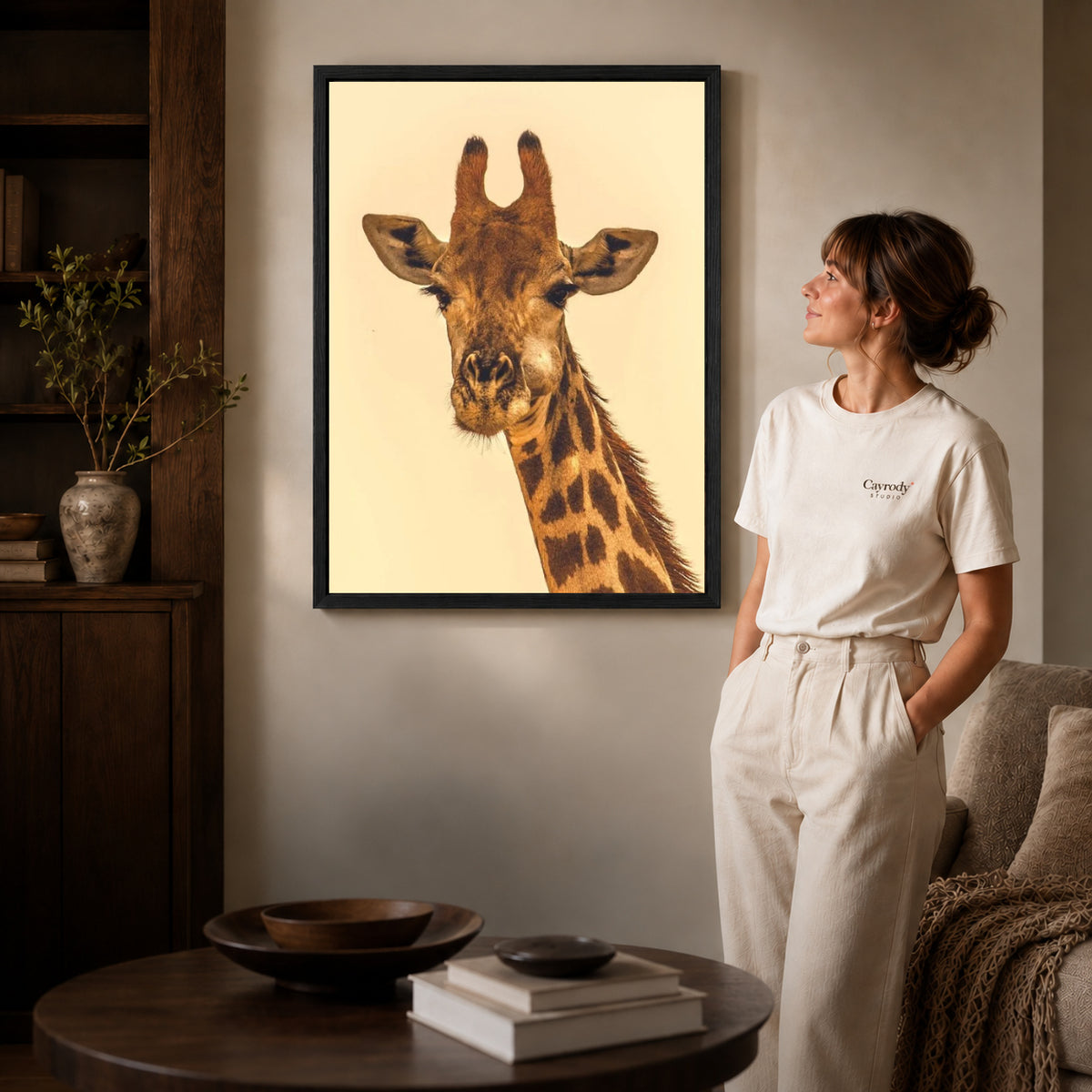 Giraffe canvas art in a black frame beside a woman and round wooden table.