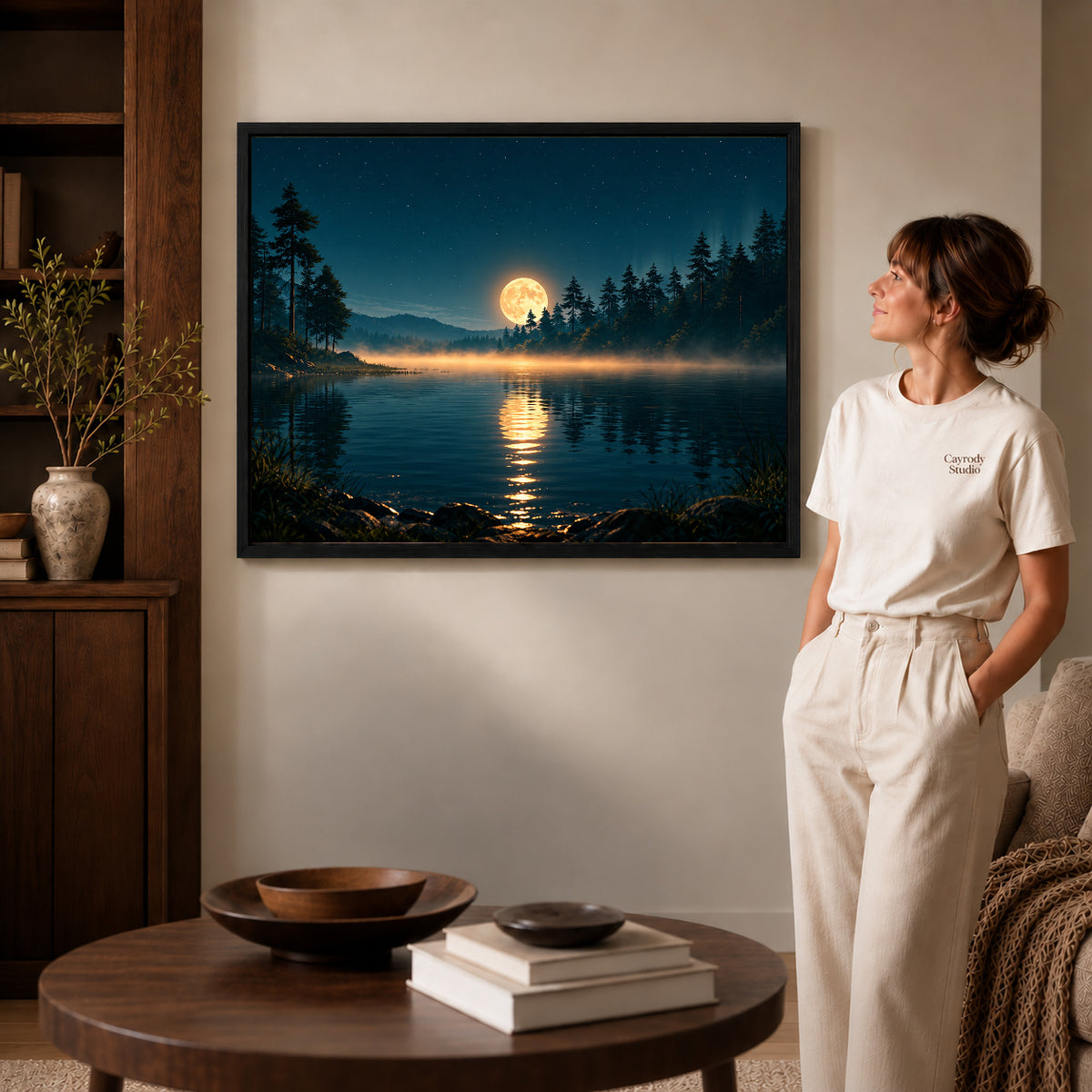Forest moon canvas art in a black frame beside a woman and round wooden table.