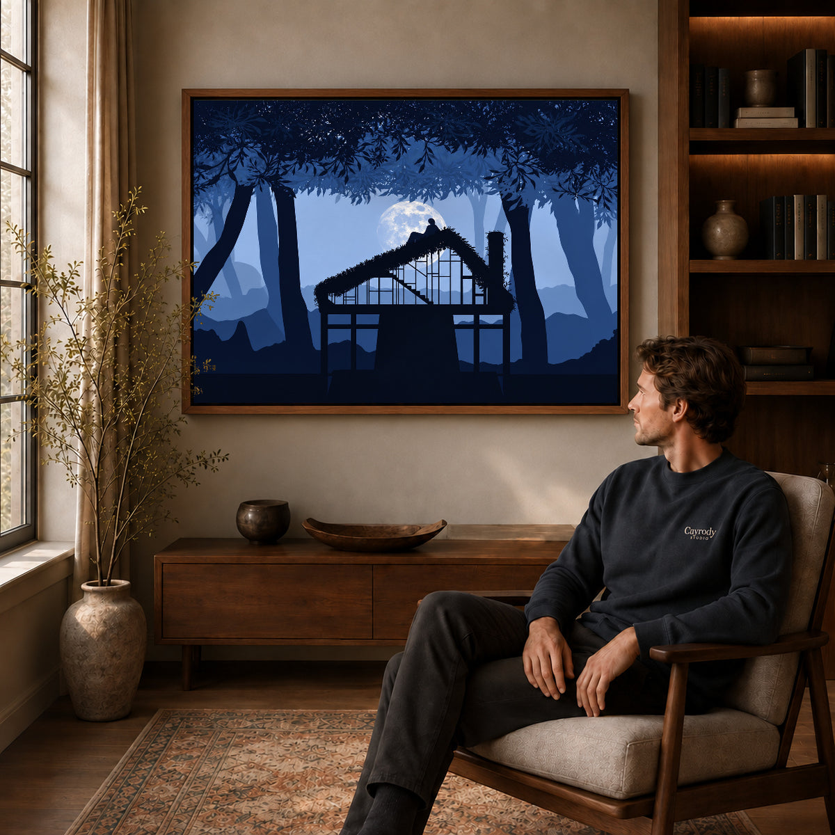 Moonlit cottage canvas art in a walnut frame above a console beside a seated man.