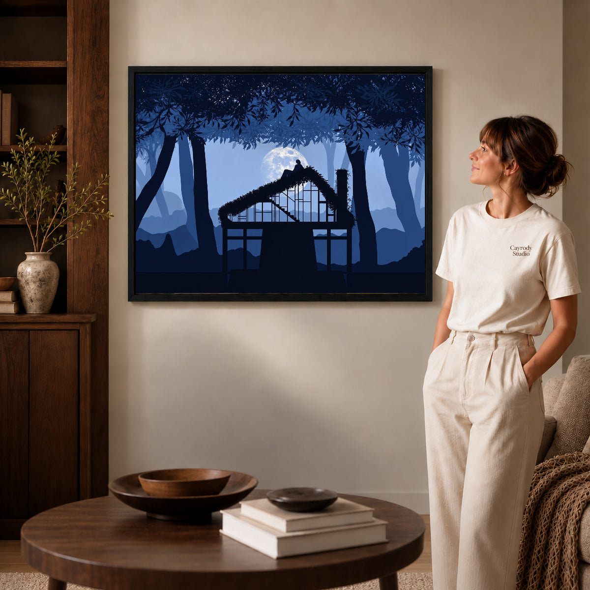 Moonlit cottage canvas art in a black frame beside a woman and round wooden table.