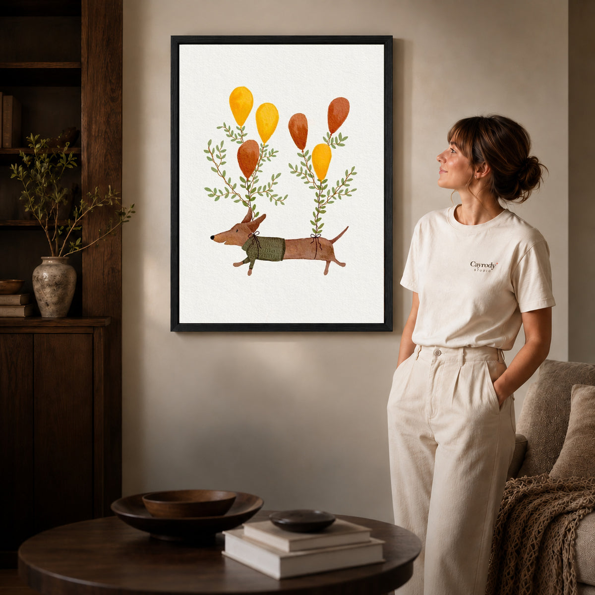 Balloon dachshund canvas art in a black frame beside a woman and round wooden table.