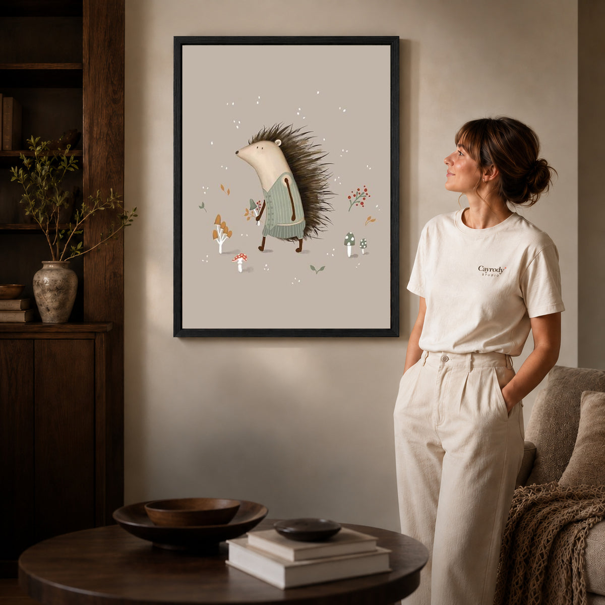 Hedgehog sweater canvas art in a black frame beside a woman and round wooden table.