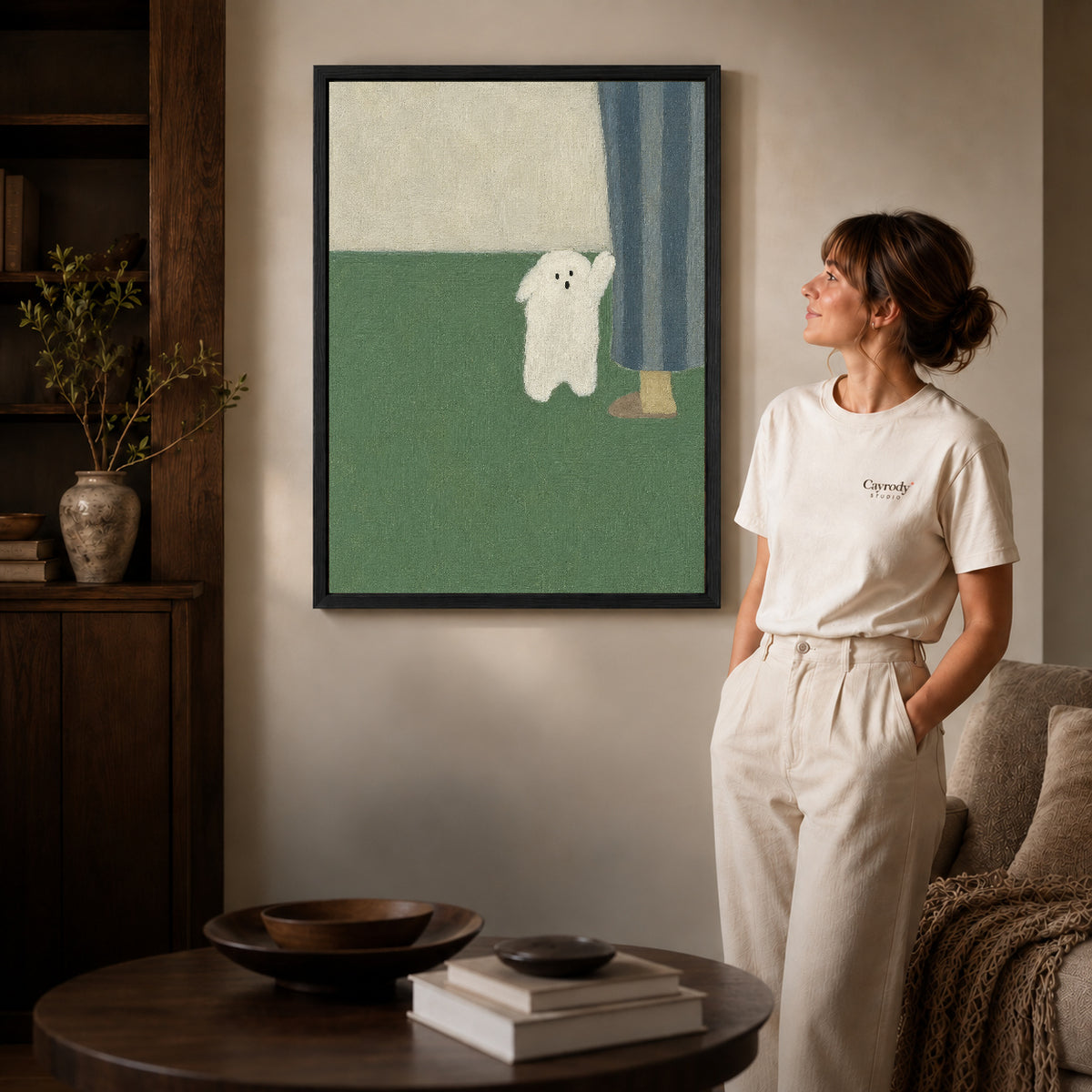 Little ghost canvas art in a black frame above a round table beside a standing woman.