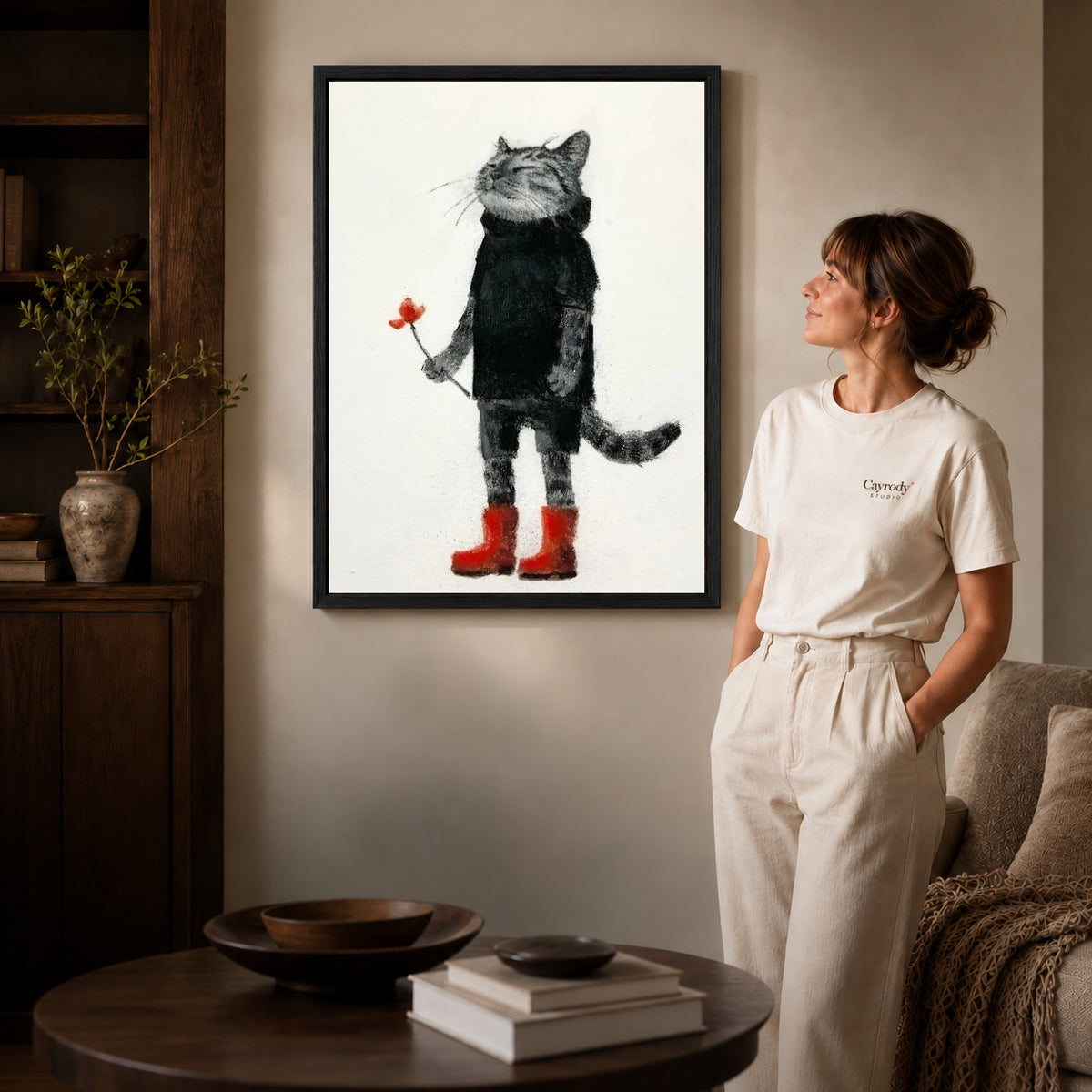 Cat red boots canvas art in a black frame beside a woman and round wooden table.