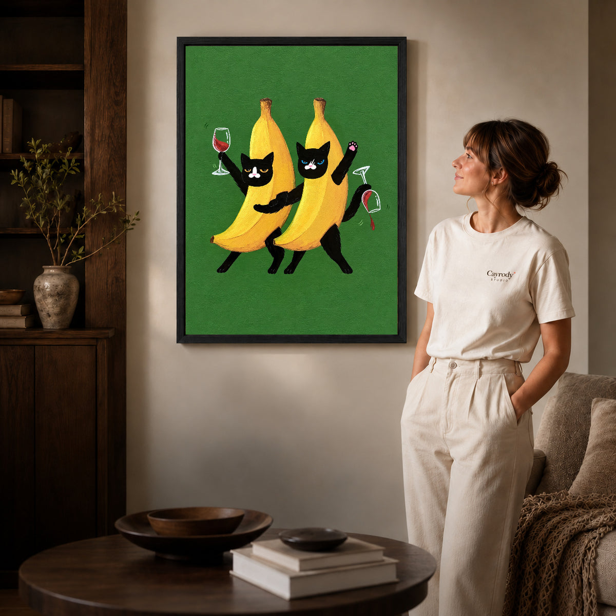 Banana cats canvas art in a black frame beside a woman and round wooden table.