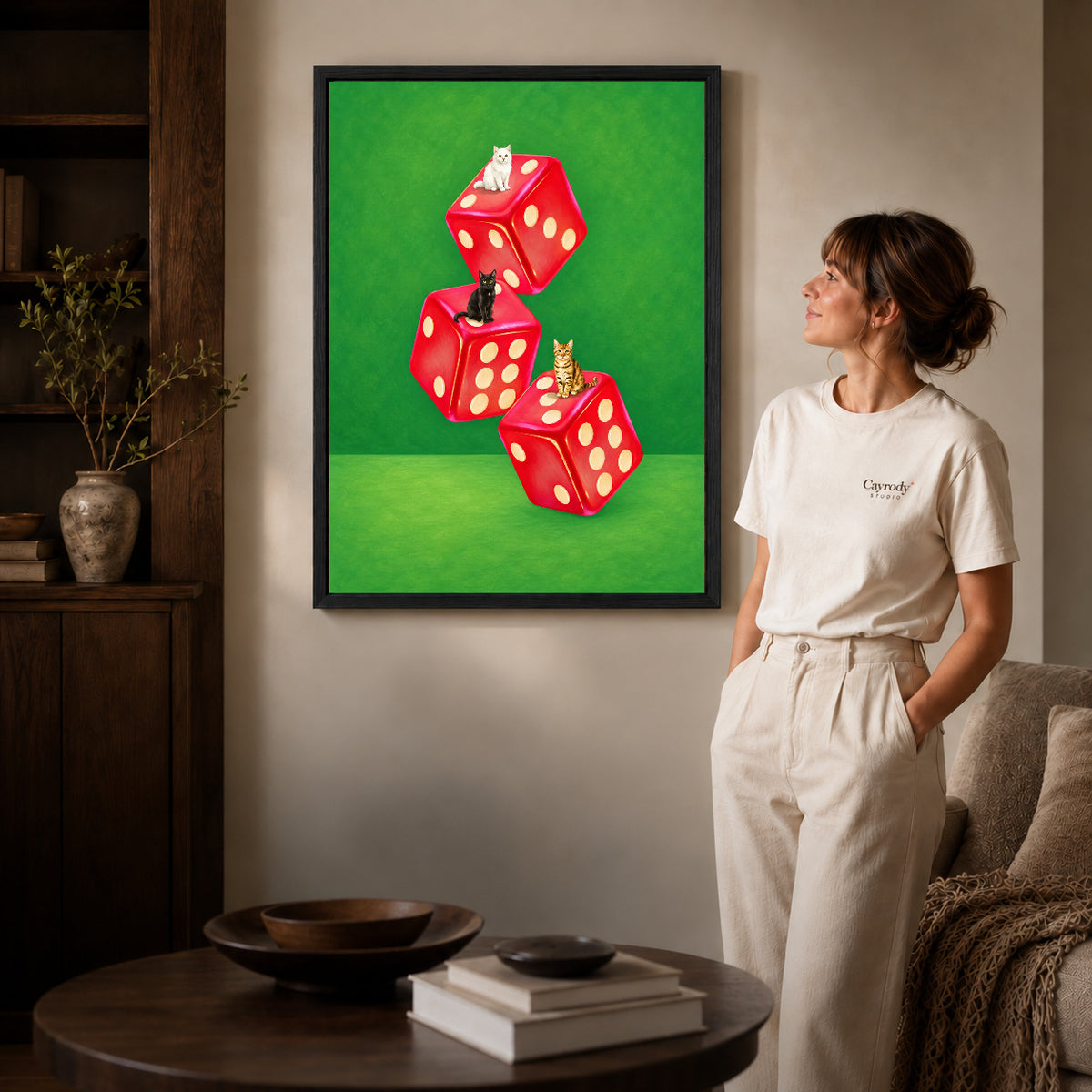 Cats and dice canvas art in a black frame beside a woman and round wooden table.