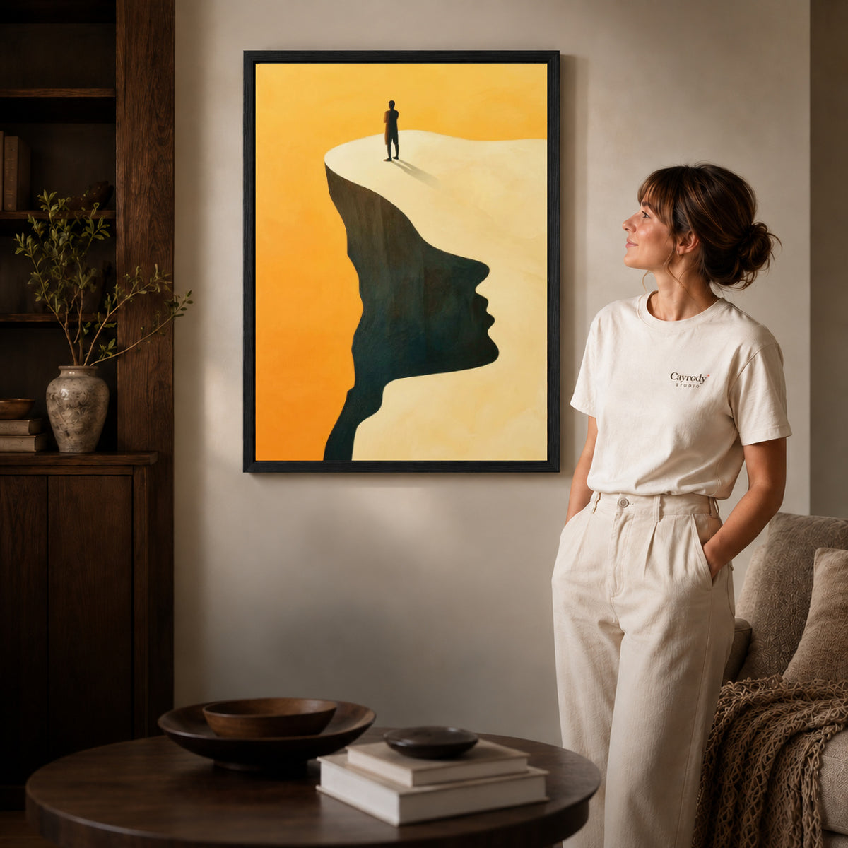 Surreal landscape canvas art in a black frame beside a woman and round wooden table.