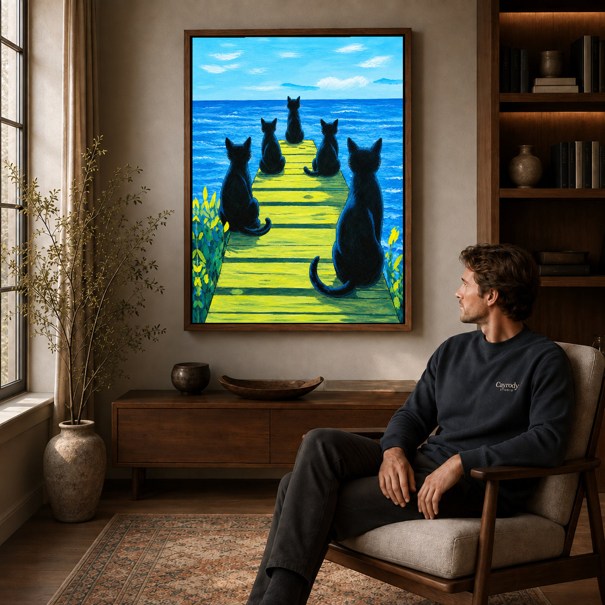 Cats ocean pier canvas art in a walnut frame above a console beside a seated man.