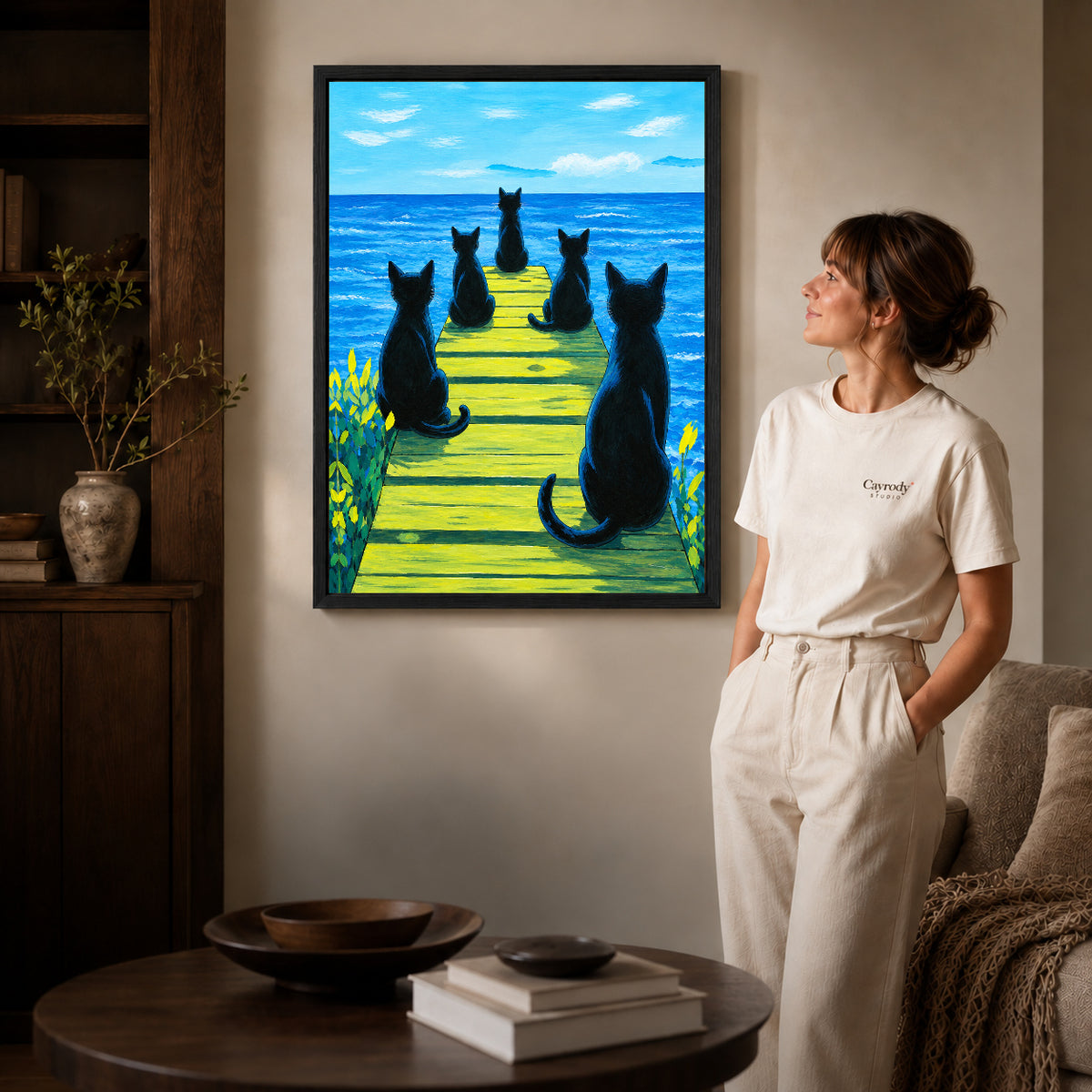 Cats ocean pier canvas art in a black frame beside a woman and round wooden table.